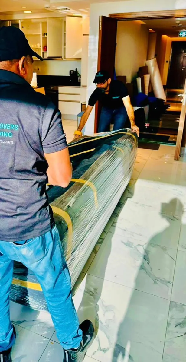Two movers carrying a sofa wrapped in blankets and stretch film through a Dubai apartment